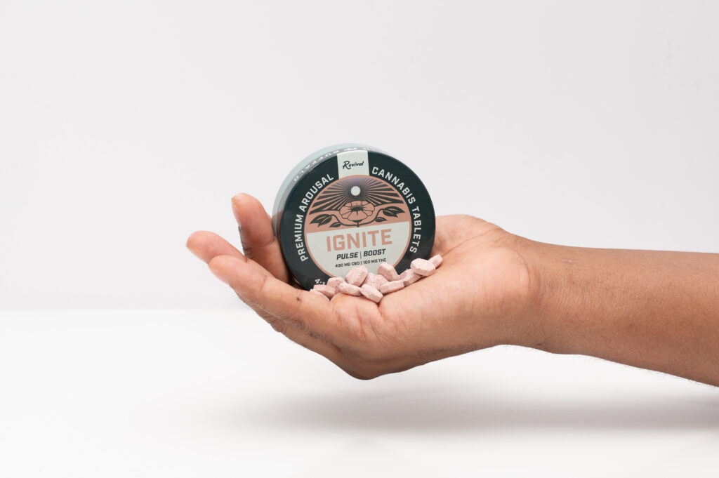 Hand holding Revival Ignite premium arousal cannabis tablets tin with pink tablets displayed in palm against a white background.