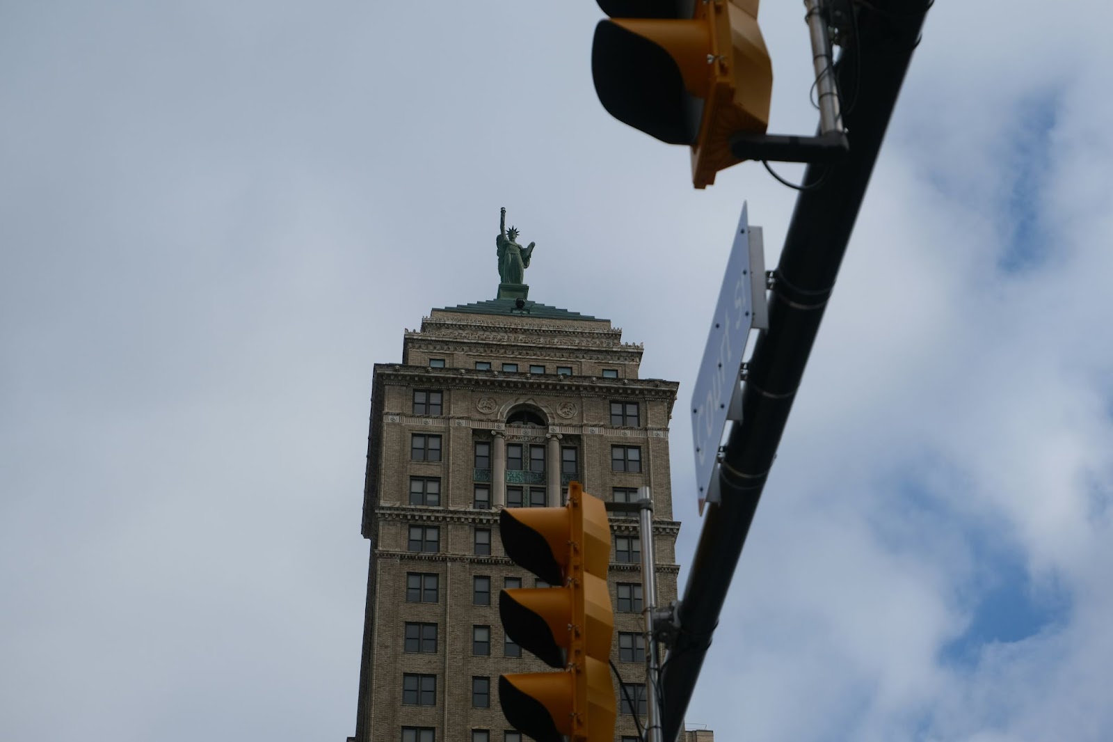 A historic high-rise building with a statue atop its roof, seen from street level in downtown Buffalo, New York, with a traffic light and street sign in the foreground against a cloudy sky