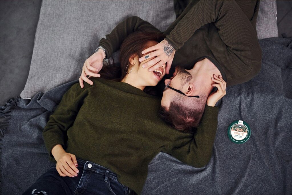 Couple lying on a cozy blanket, laughing and cuddling in an intimate moment. A tin of Revival Gems “IGNITE” cannabis tablets is placed nearby, highlighting the product’s connection to mood enhancement.