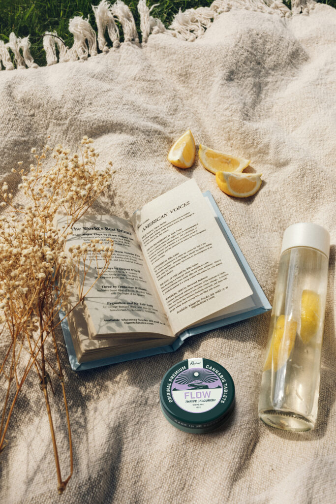 Flat lay of a peaceful outdoor setup featuring a book open to a poem, a water bottle with lemon slices, dried wildflowers, and a tin of Revival Gems “FLOW” cannabis tablets placed on a soft beige blanket.