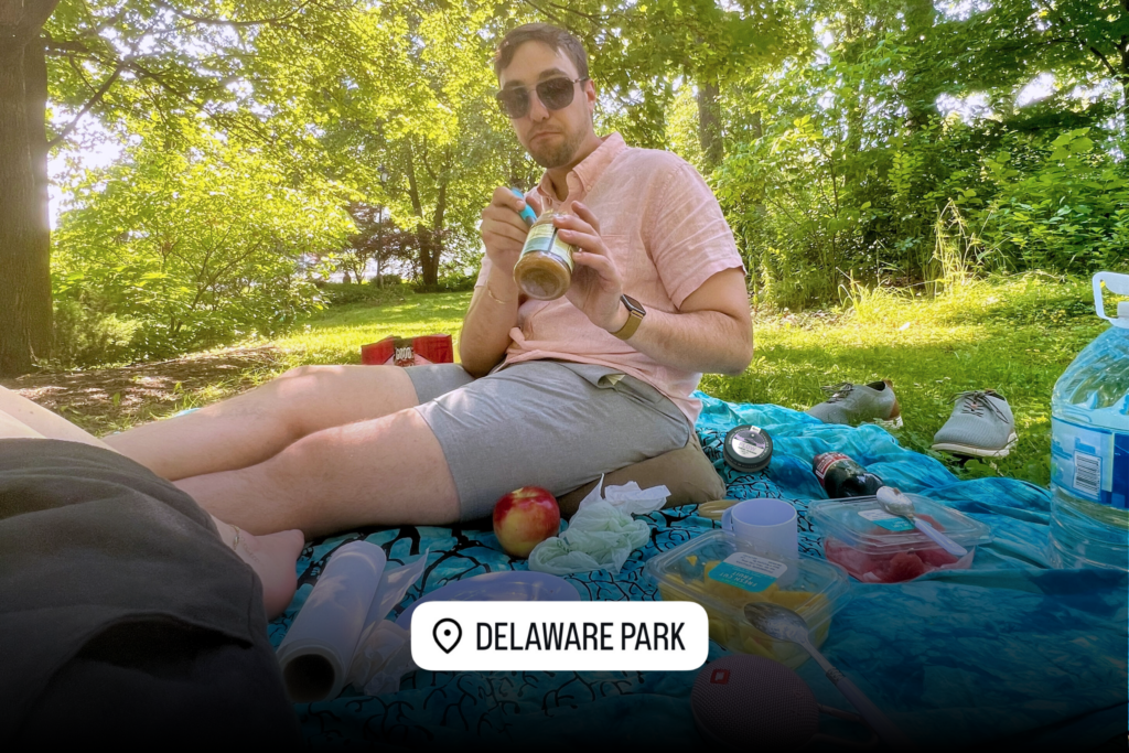 A man sits on a picnic blanket at Delaware Park, surrounded by snacks and picnic supplies. A tin of Revival Gems Flow cannabis tablets is visible on the blanket near him, along with an apple, plastic containers, and a bottle of soda. Sunlight filters through the trees in the background.