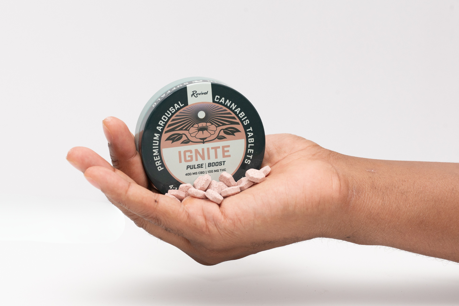 A hand holds a tin of Revival Gems Ignite cannabis tablets, clearly labeled “Ignite – Pulse | Boost.” Several light orange hexagonal tablets spill into the palm. The label highlights 400 mg CBD and 100 mg THC. Shot against a white background, the image emphasizes the product’s compact size and on-the-go usability.