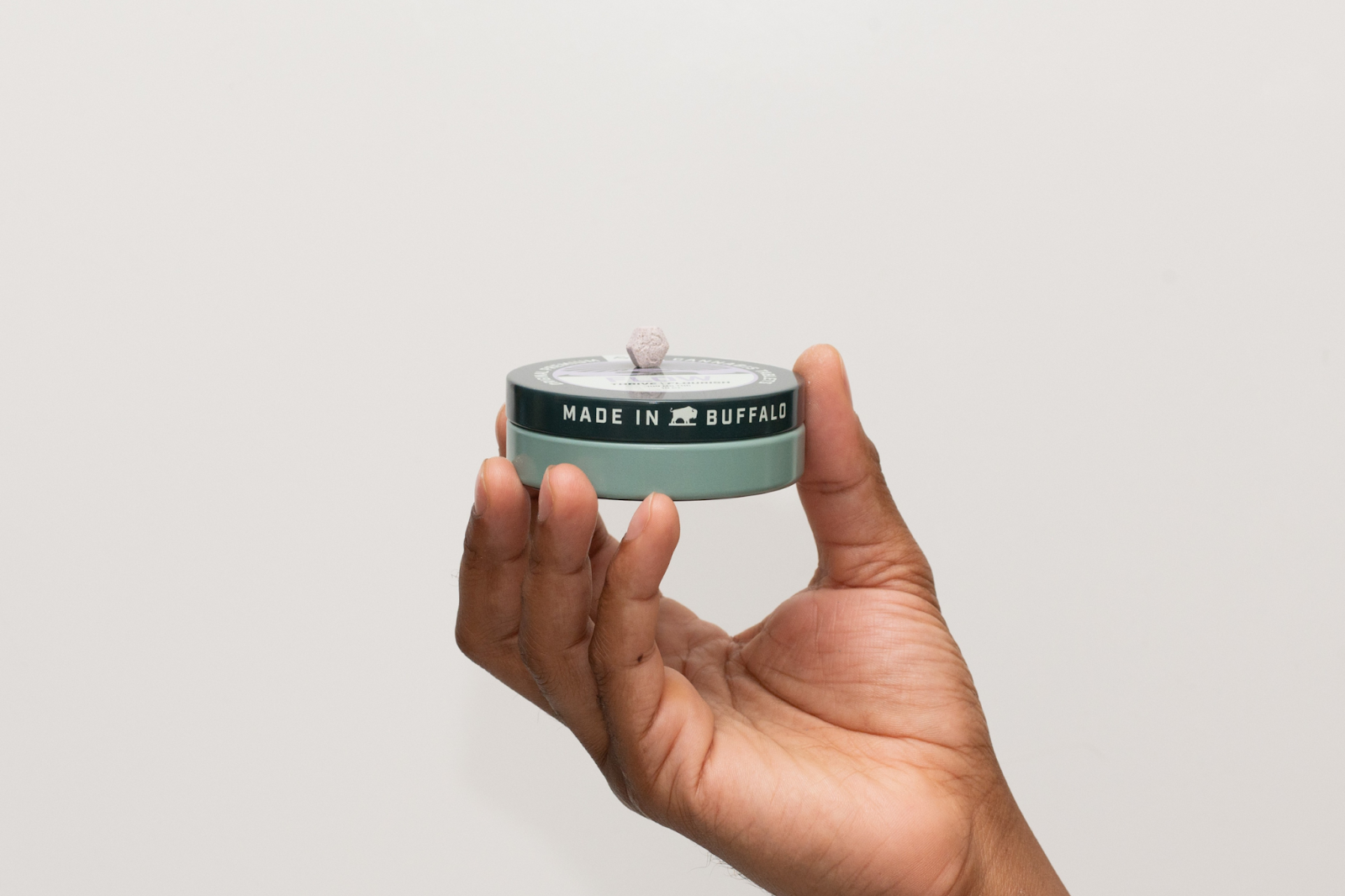 A hand holds a tin of Revival Gems Drift cannabis tablets, labeled “Made in Buffalo.” One blue hexagonal tablet is balanced on top of the teal and green tin. The product features a 2:1:1 blend of CBN, THC, and CBD, formulated for sleep and nighttime relaxation. Set against a plain white background.