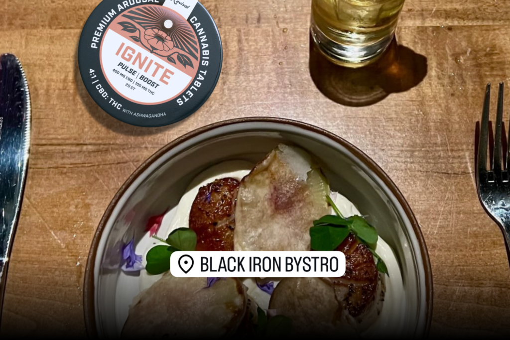 A dinner table at Buffalo restaurant Black Iron Bystro set for a date night, featuring a plated dish with seared scallops and garnishes, a drink, and silverware. A tin of Revival Gems Ignite cannabis tablets is placed on the table in the upper left corner.

