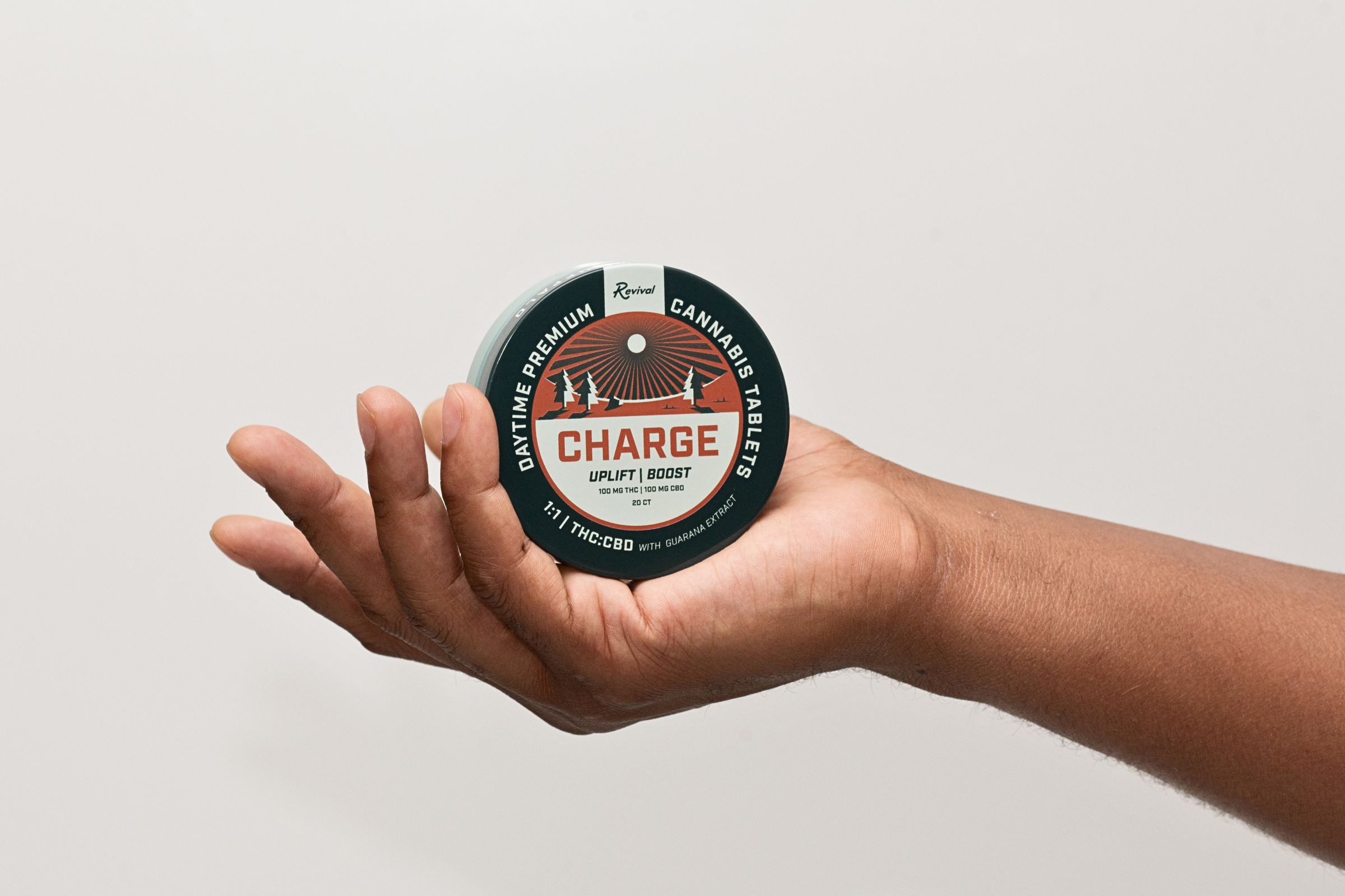 A hand holding a round container of Revival Charge cannabis tablets. The label reads "Daytime Premium Cannabis Tablets – Charge – Uplift | Boost," with 100 mg THC and 100 mg CBD. The design features trees and a rising sun, and the label notes a 1:1 THC:CBD ratio with guarana extract.
