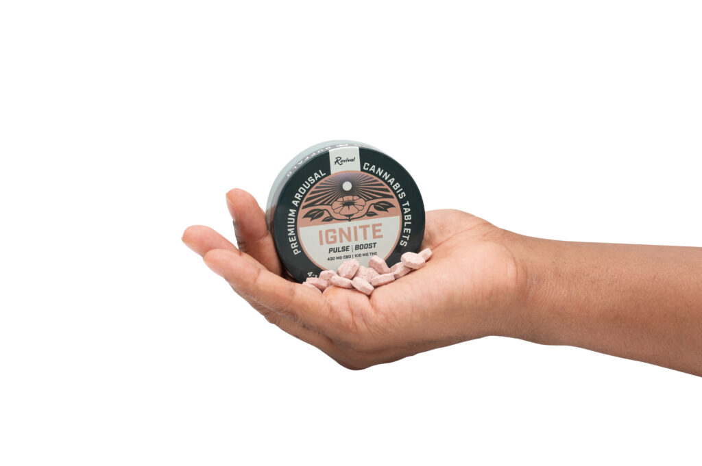 Hand holding a tin of Revival Gems ‘Ignite’ cannabis tablets with several pink, hexagon-shaped tablets spilling out — a CBD-forward formula crafted for intimacy, relaxation, and connection. Designed by healthcare professionals, vegan and full-spectrum.