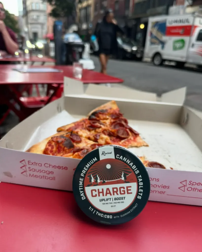 Revival “Charge” daytime premium cannabis tablets (1:1 THC:CBD with guarana extract) placed on a red outdoor café table in front of a pizza box with slices of pepperoni pizza. The label reads “Uplift | Boost,” and the city street scene is softly blurred in the background. Revival is a locally made brand in Buffalo, NY.