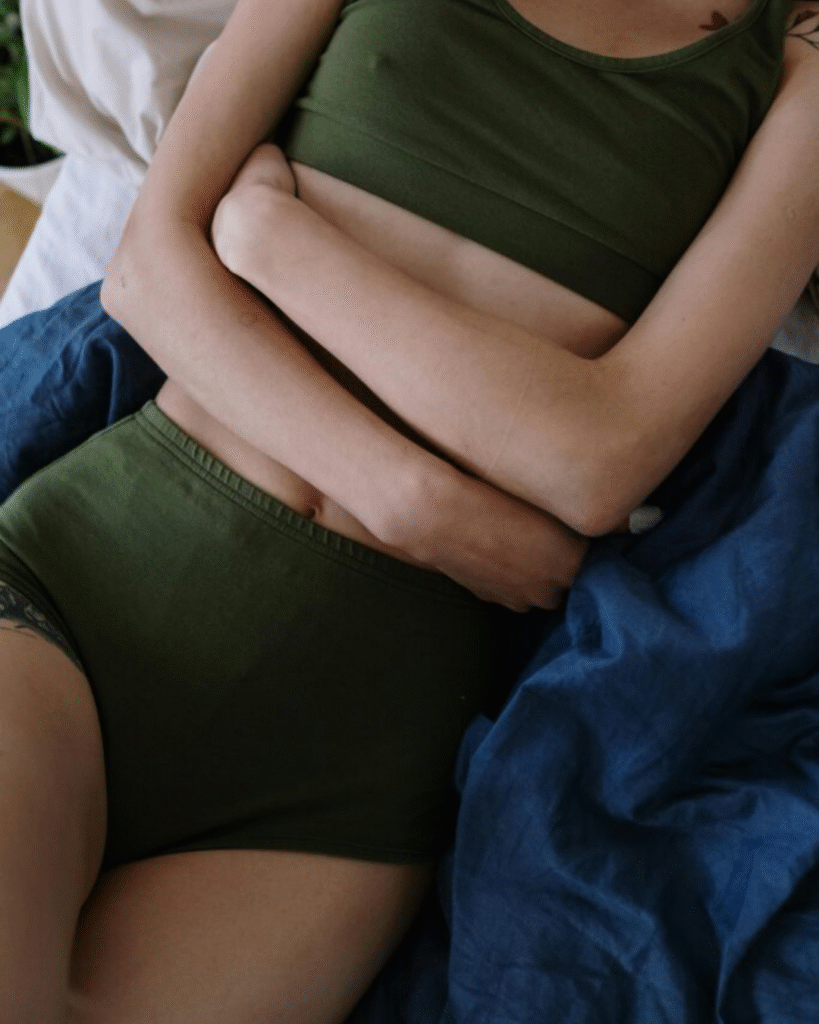 Person lying on a bed in green loungewear, arms wrapped around their lower abdomen in a protective posture, visually representing pelvic pain and discomfort associated with endometriosis while referencing symptom relief support from Revival Gems.