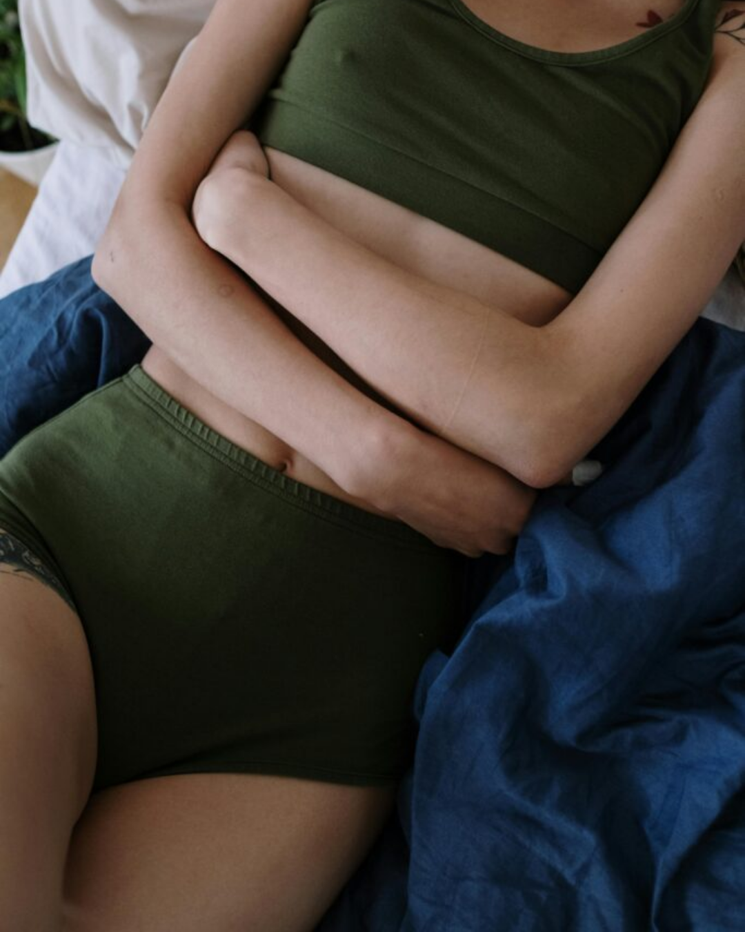 Person lying on a bed in green loungewear, arms wrapped around their lower abdomen in a protective posture, visually representing pelvic pain and discomfort associated with endometriosis while referencing symptom relief support from Revival Gems.
