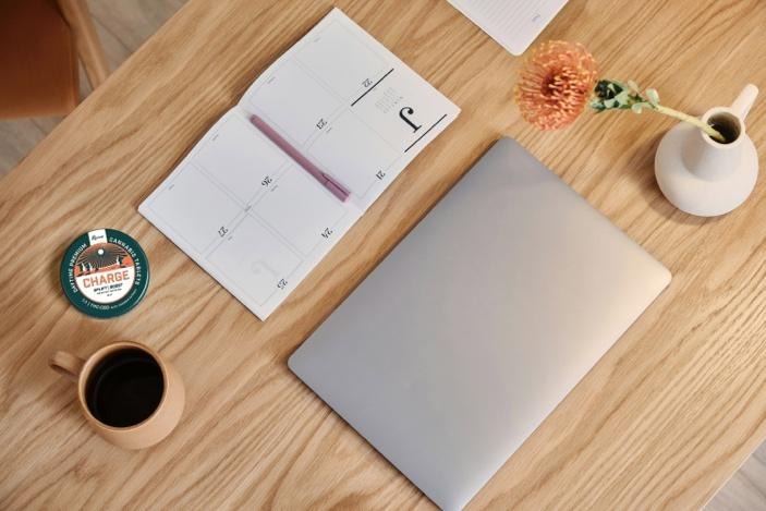 Wooden desk with a closed laptop, open planner, coffee cup, small vase with a flower, and a tin labeled Charge energy tablets.