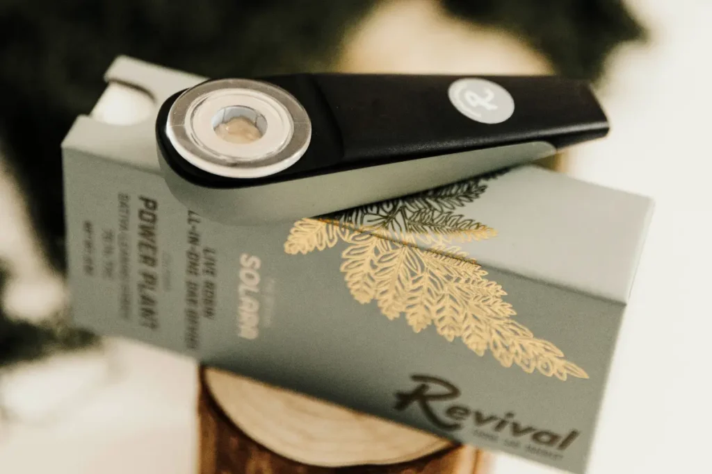 Close-up of Revival’s Solara live rosin dab device in black and sage green, showing the visible live rosin concentrate inside the chamber. The device rests on green and gold Solara packaging labeled “Power Plant” (sativa-leaning hybrid) and “live rosin all-in-one dab device.” Revival is a locally made brand in Buffalo, NY. Flavor: Solara live rosin – Power Plant.