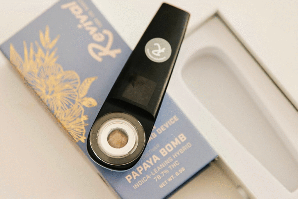 Close-up of a Revival's Solara: A pocketable rosin dab device displaying the live rosin inside, placed on blue and gold packaging labeled “Papaya Bomb” (indica-leaning hybrid). Revival is a locally made brand in Buffalo, NY. Flavor: Papaya Bomb live rosin.