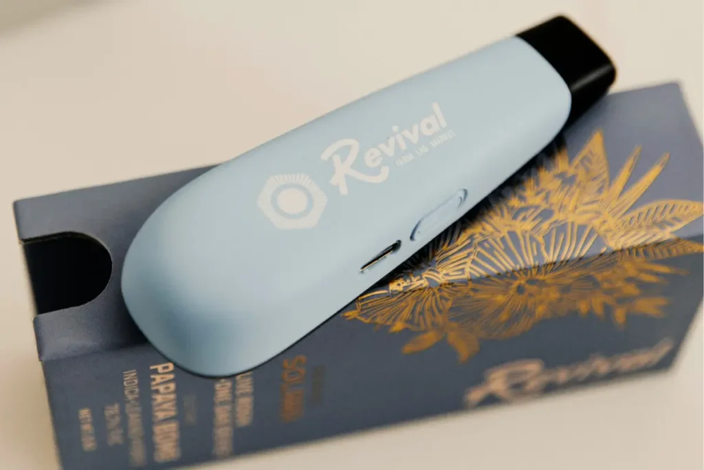 Light blue Revival Farm Lab Market Solara pocketable live rosin dab device resting on blue and gold “Papaya Bomb” (indica-leaning hybrid) packaging. The device features a matte finish with a side button and charging port visible. Revival is a locally made brand in Buffalo, NY. Flavor: Papaya Bomb live rosin.